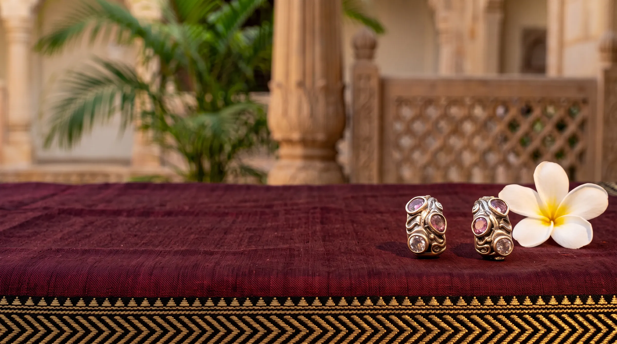 Silver earrings set with pink stones on maroon handloom beside a frangipani