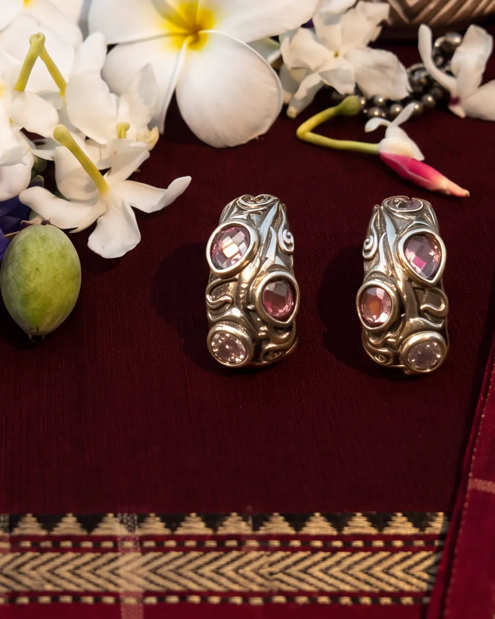 Silver earrings set with pink stones on maroon handloom beneath frangipani and butterfly-pea flowers