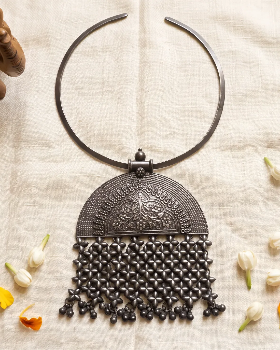 A silver hasli necklace laid on ivory linen with jasmine buds and a carved wooden elephant